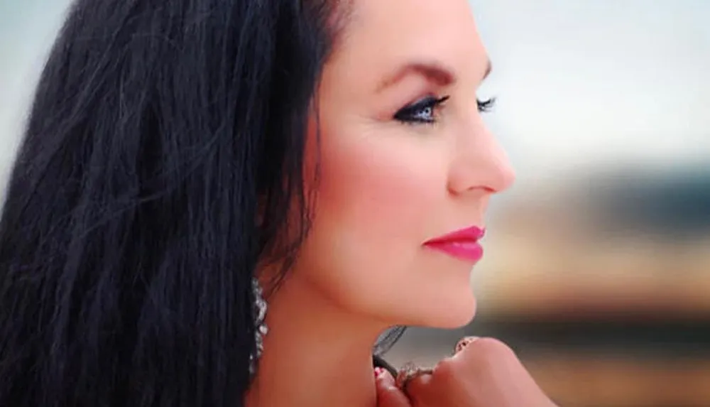 The image shows a close-up profile of a woman with dark hair pink lipstick and prominent eye makeup gazing off to the side against a blurred background
