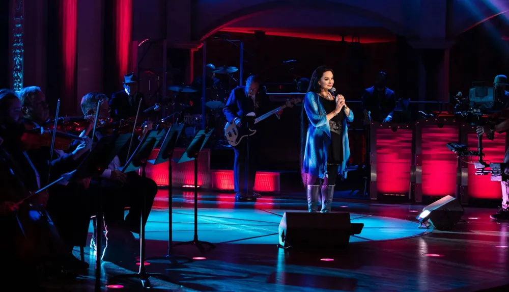 A singer performs onstage with a live orchestra and band in a concert setting with warm stage lighting