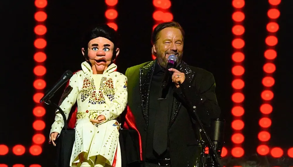 A ventriloquist performs on stage with a puppet dressed in a bejeweled jumpsuit