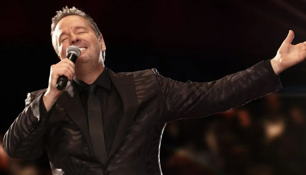 A man in a patterned suit passionately sings into a microphone with one arm outstretched