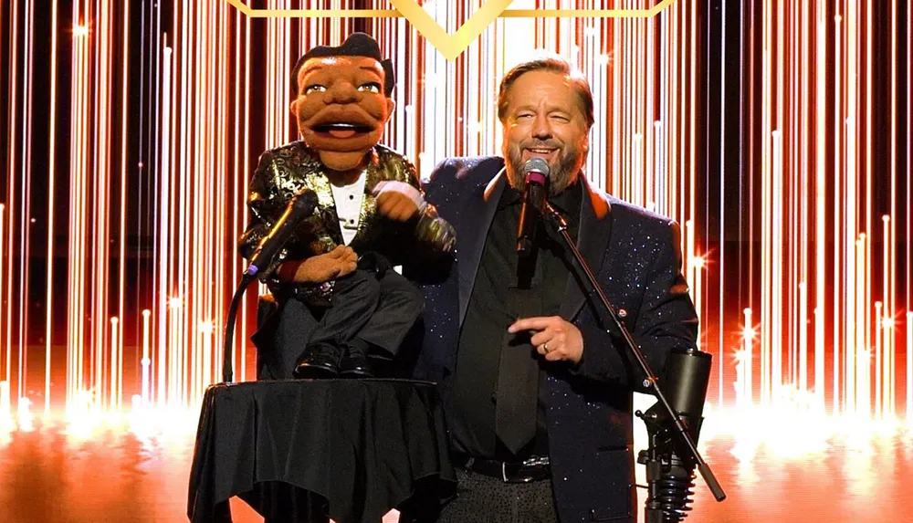 A ventriloquist performs on stage with a puppet against a backdrop of vertical light streaks
