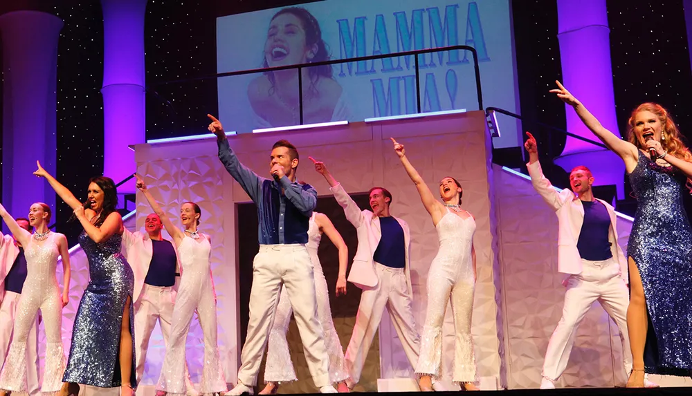 Performers in sparkling costumes are singing and dancing on stage during a production that appears to be themed around the musical Mamma Mia based on the screen projection and attire