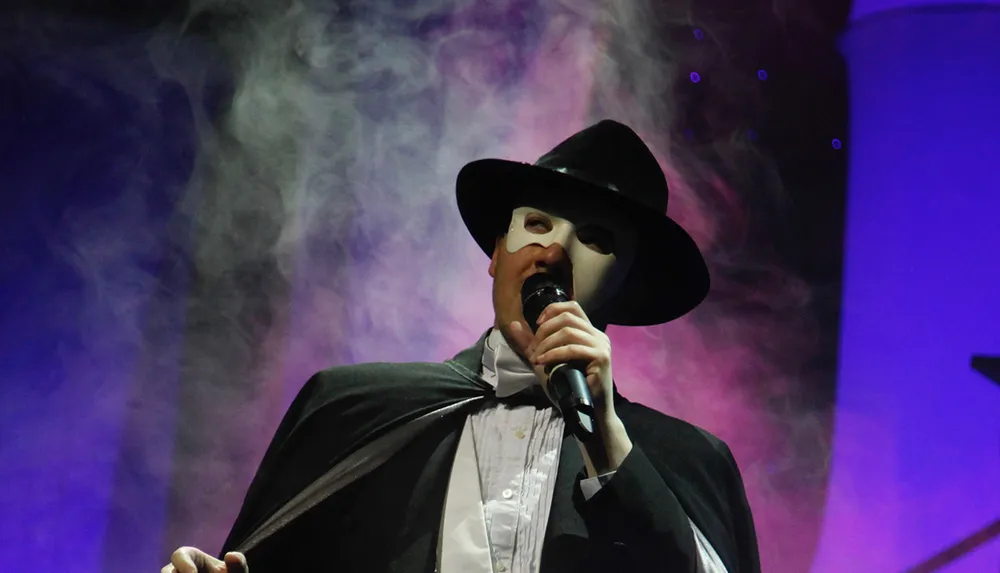 A person wearing a mask and a black hat is performing on stage with a microphone surrounded by colorful stage lighting and smoke