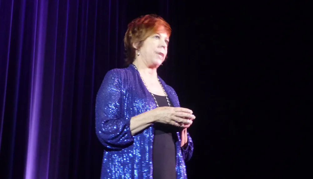 A person in a sparkly blue jacket is standing on stage with a microphone likely addressing an audience