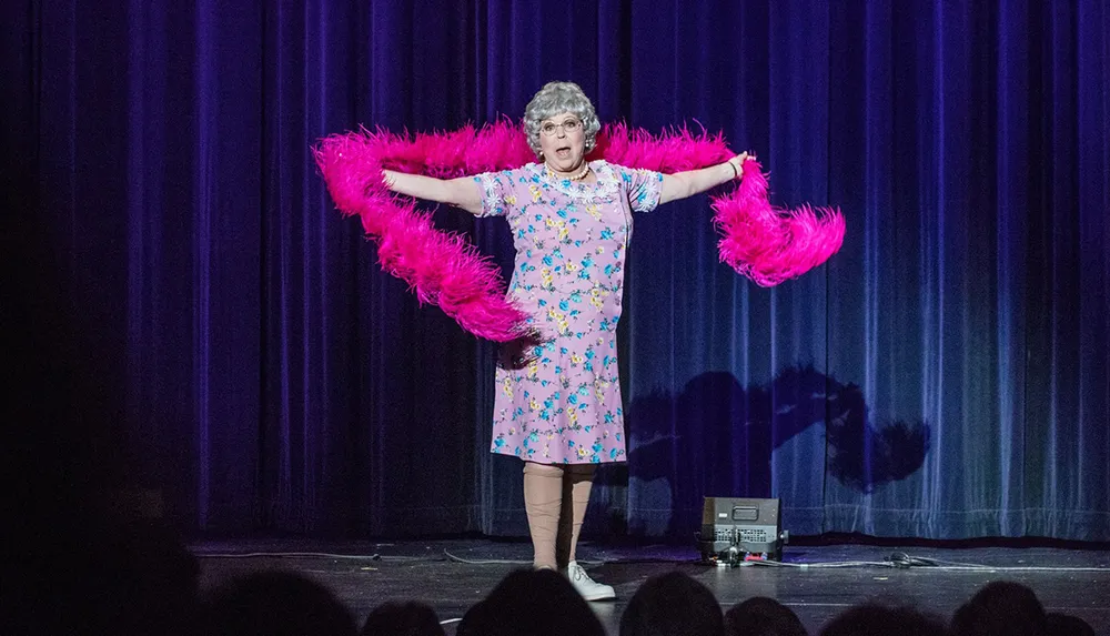 A performer in a floral dress and glasses is holding pink feather boas with arms outstretched on a stage with a dark curtain backdrop