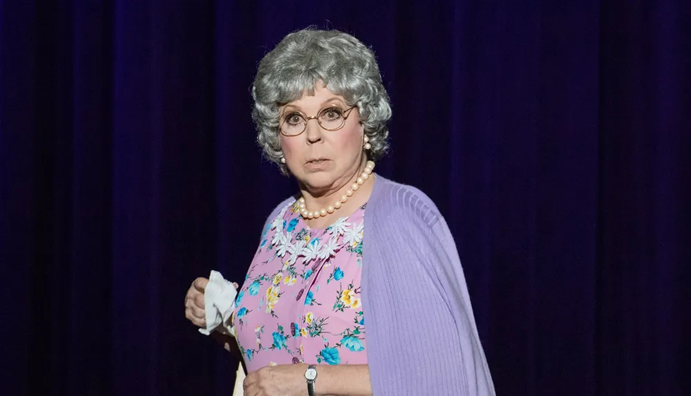 A person is standing on a stage wearing a lilac cardigan over a floral dress glasses a pearl necklace and a gray hairstyle with a surprised or concerned expression