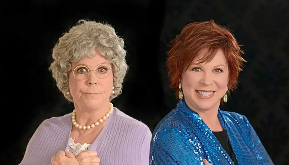 Two women one portraying an older character with gray hair glasses and a purple sweater and the other with reddish hair and a blue sequined jacket are side by side against a black background