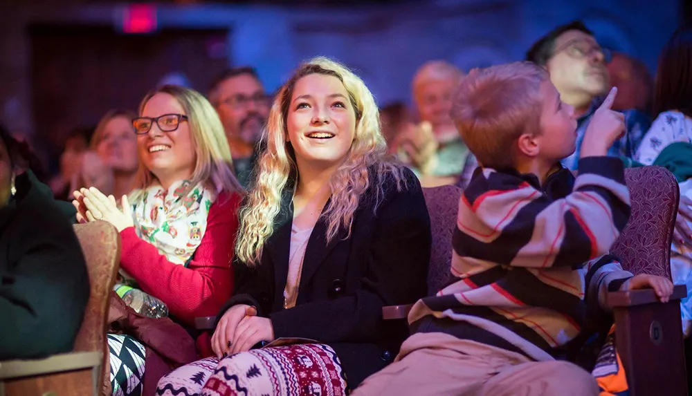 An audience is watching a performance appearing engaged and delighted with a young woman in the foreground smiling and clapping