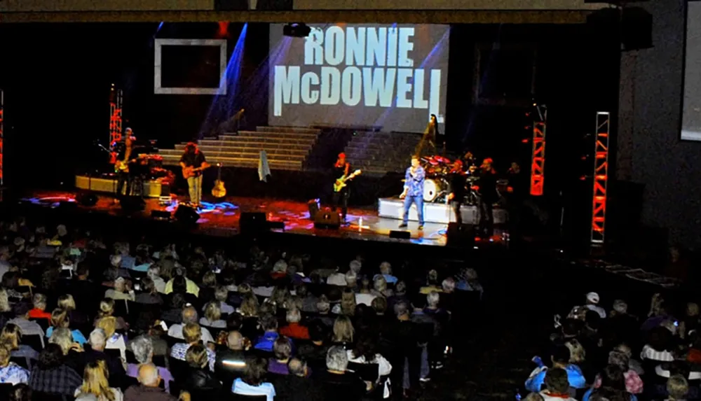 A live concert is taking place in front of an audience with the name Ronnie McDowell projected in large letters on the screen behind the stage